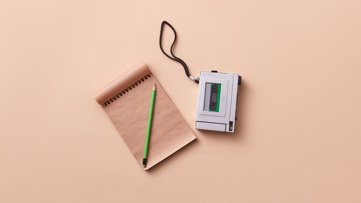 Top view of a vintage dictaphone, spiral notebook, and green pencil on a beige background.